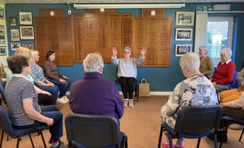 A group of people aged 70+ sat with their hands in the air doing seated exercising