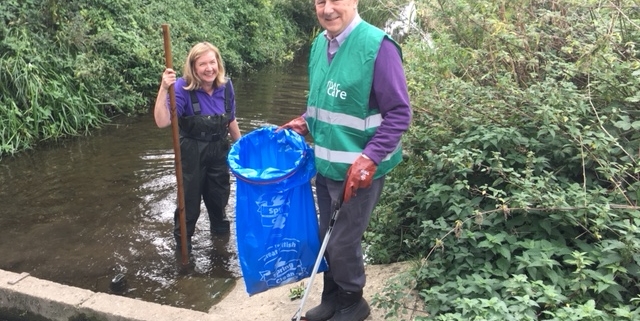 A blond lady wearing waders in the river with a gentleman holding a blue bin bag on the bank
