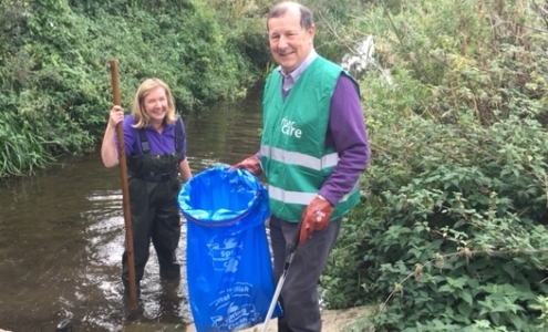 A blond lady wearing waders in the river with a gentleman holding a blue bin bag on the bank