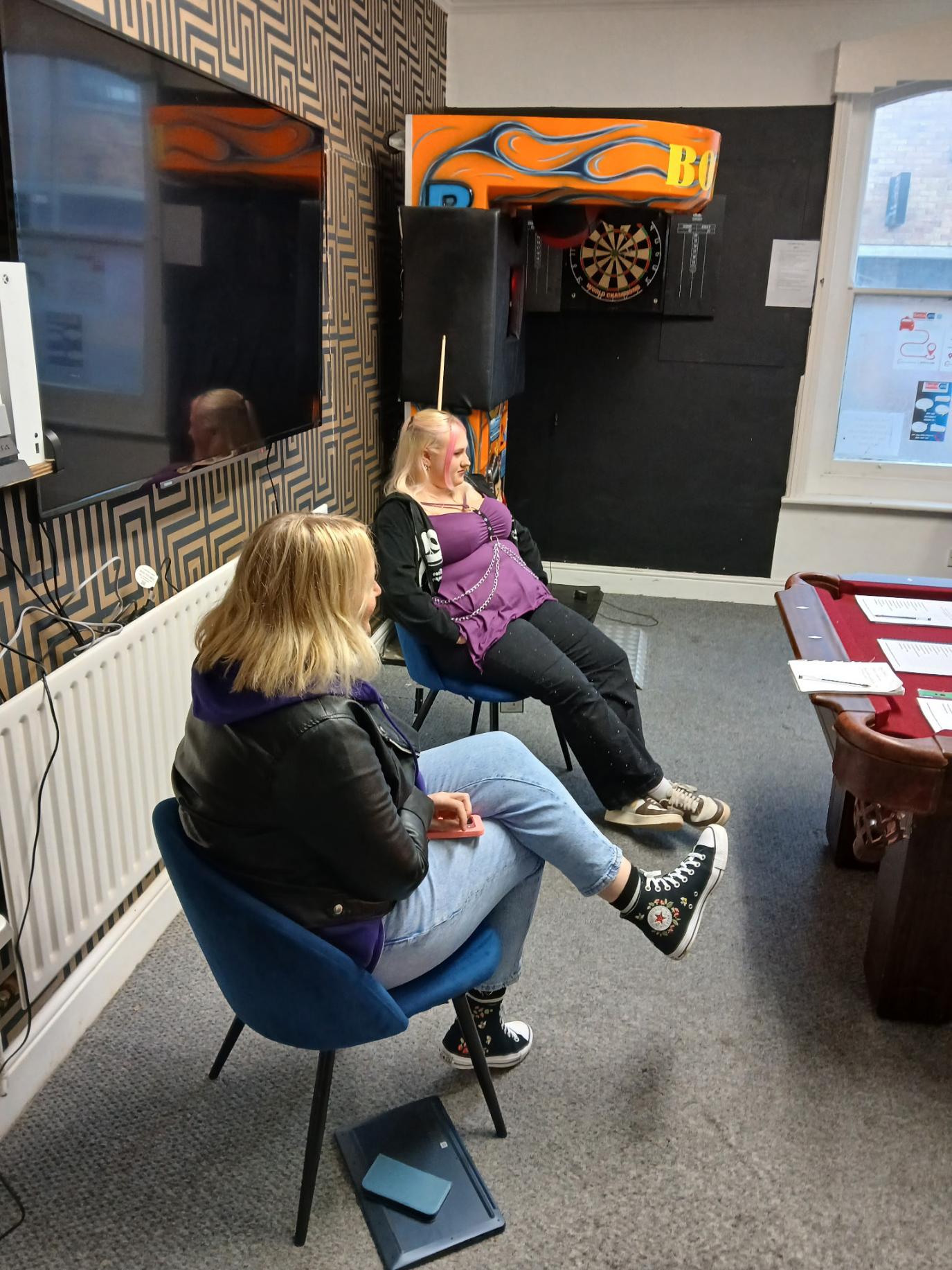 two teenage girls with blond hair sat under a TV and looking at a snooker table