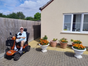 A gentleman in a mobility scooter outside a cream bungalow with lots of pots of plants out the front